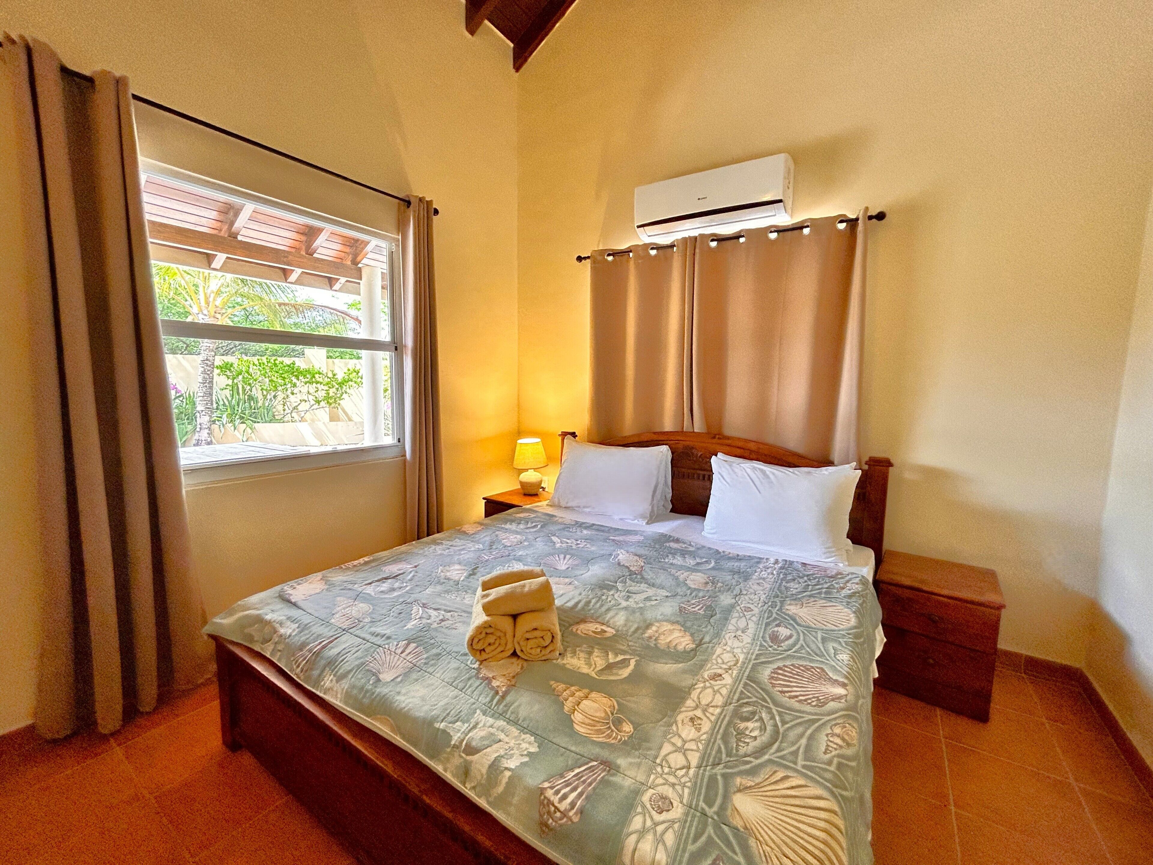 A hotel room with a bed, a nightstand, a lamp, a window with curtains, and an air conditioning unit.