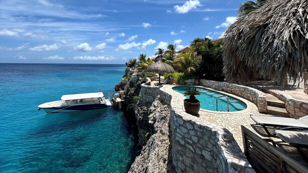 A clear blue sea, a boat docked at a stone pier, a pool surrounded by stone walls, and a thatched-roof structure. - Lagun Blou Resort (Lagun)