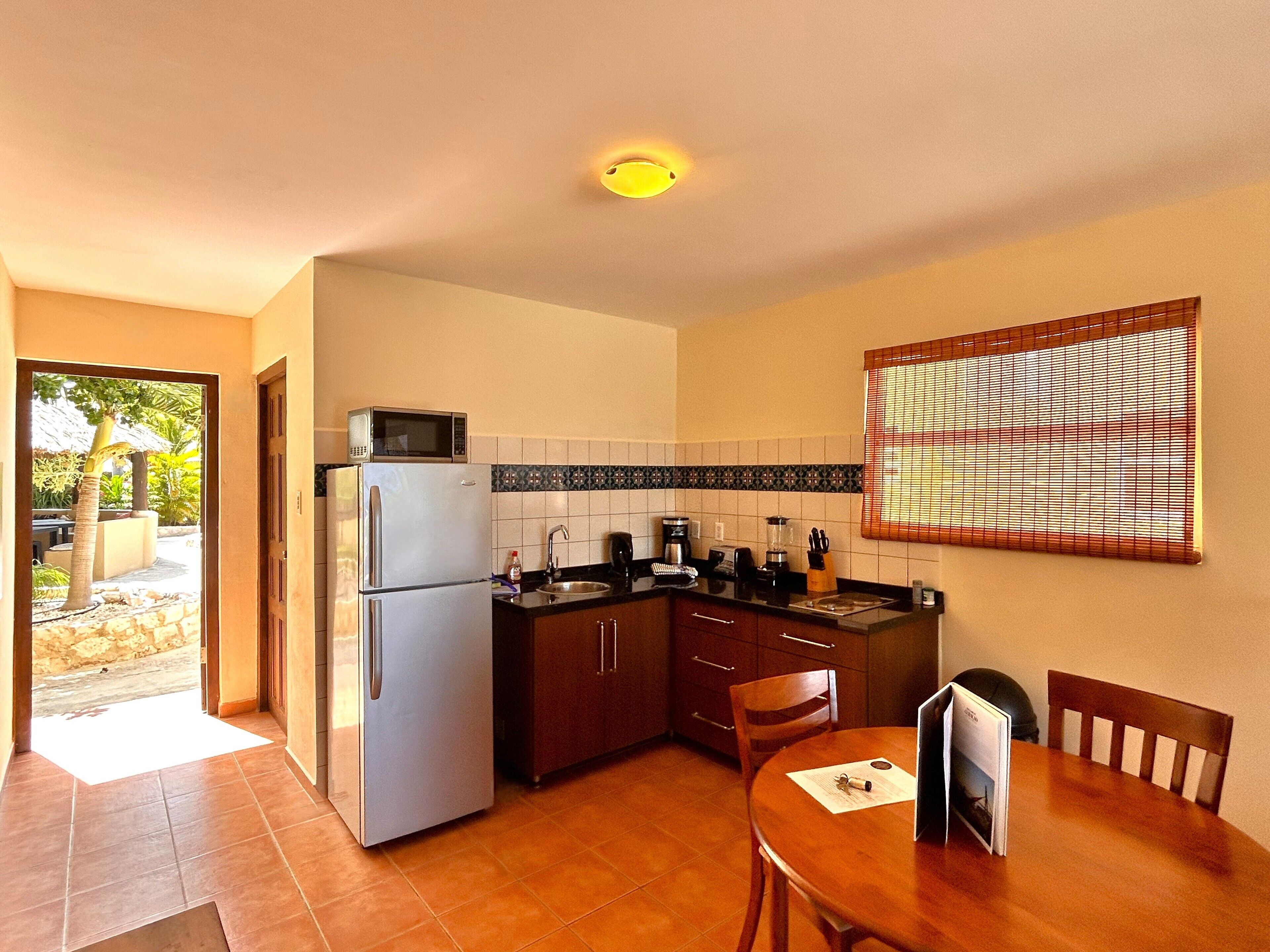 Una cocina compacta con refrigerador, microondas y cafetera. Hay una mesa de comedor con sillas y una ventana con persianas.