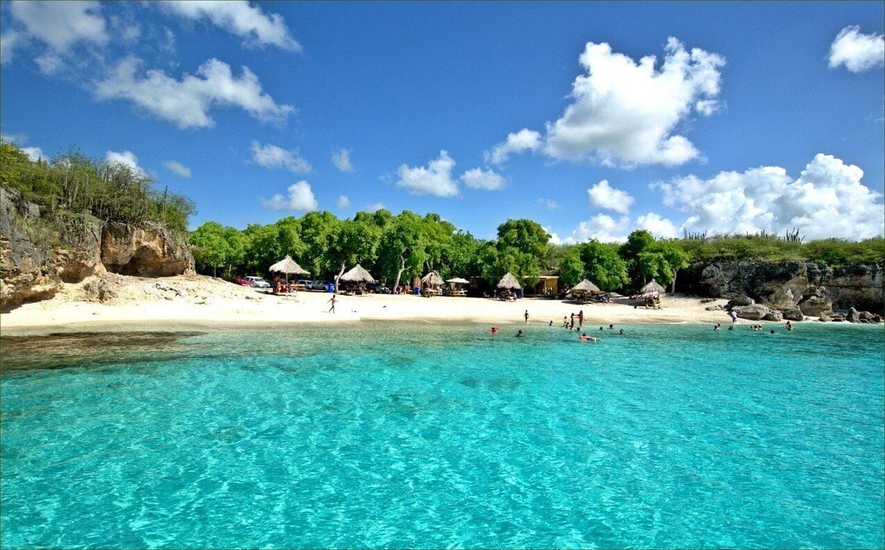 A beach with clear turquoise water, white sand, and thatched-roof huts.