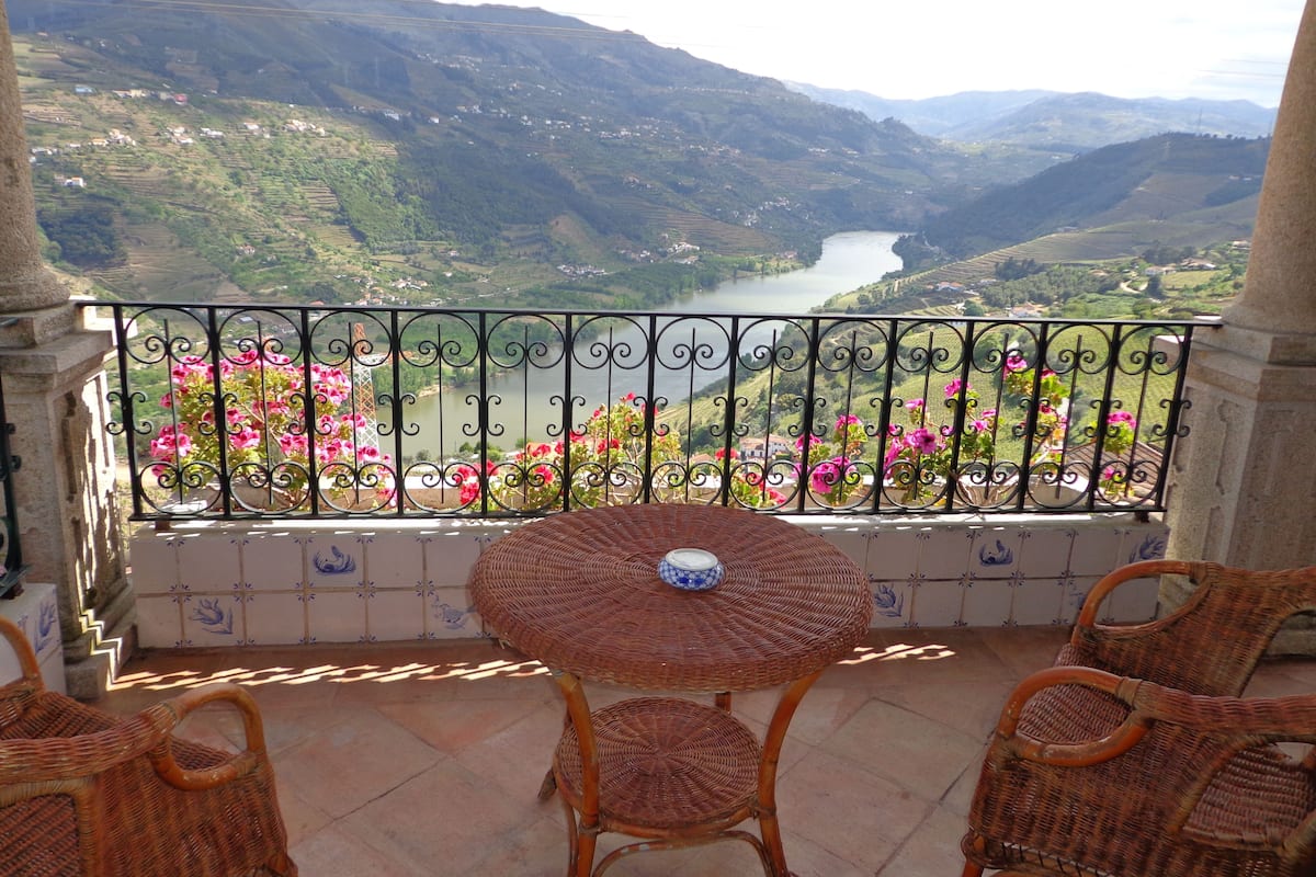 A balcony with wicker chairs, a table, and a view of a river and mountains.