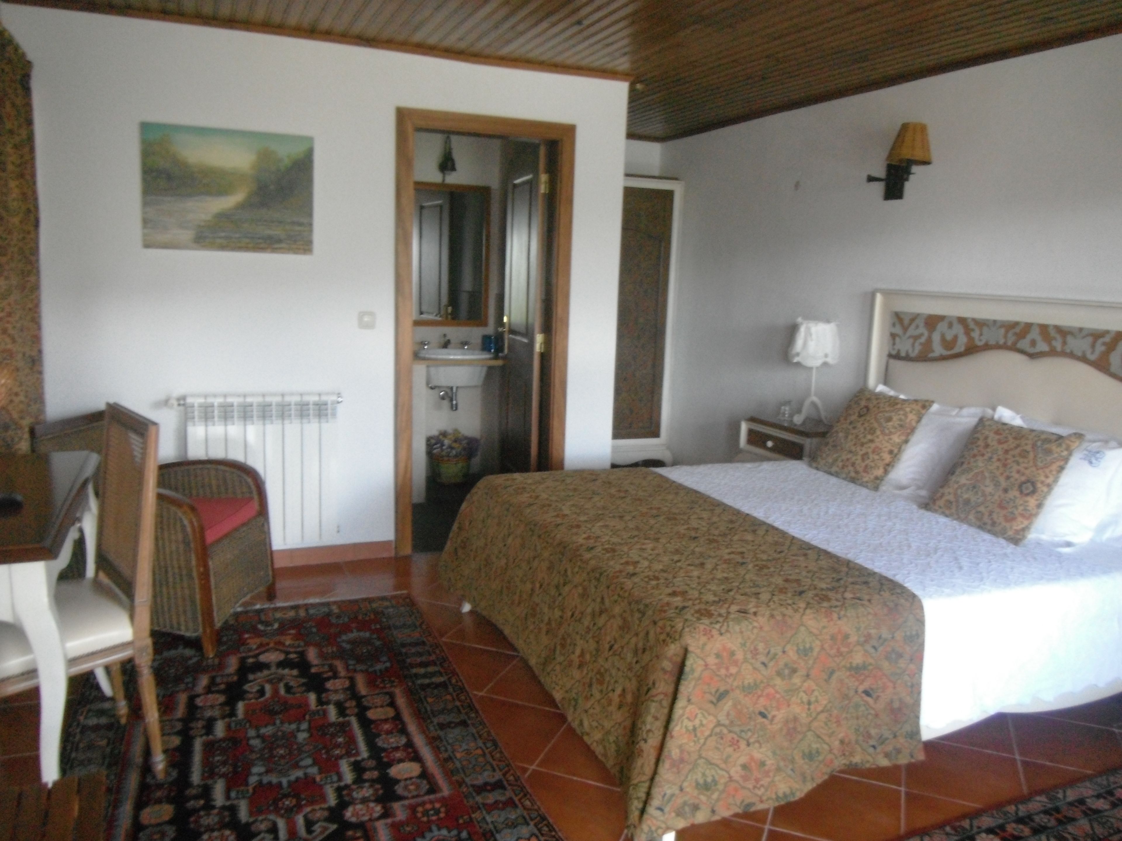 A bedroom with a bed, a nightstand, a lamp, a dresser, a chair, a radiator, and a door leading to a bathroom.