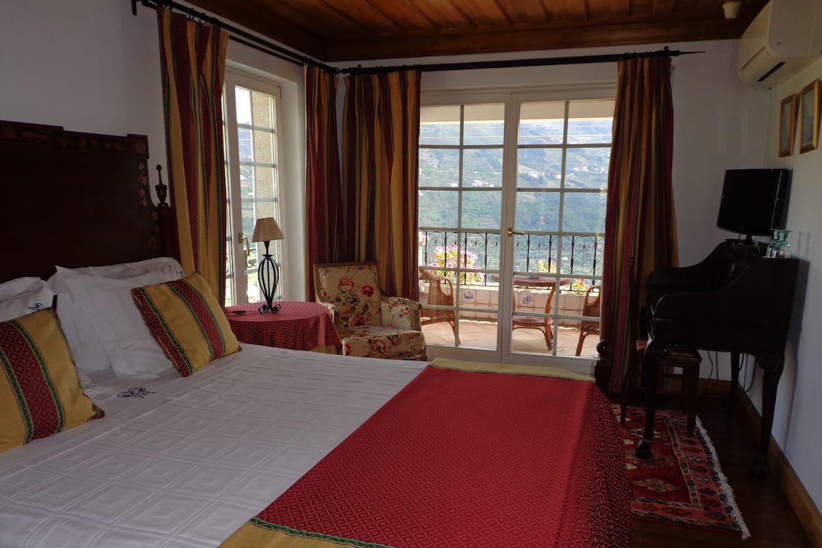 A hotel room with a large bed, a piano, a balcony with chairs, and a view of the countryside.
