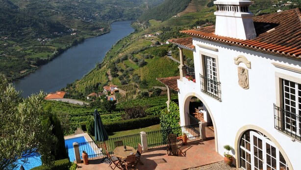 A white building with a tiled roof overlooking a river and lush green hills. - Casa de Canilhas (Mesão Frio)