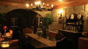 A dimly lit dining area with a long table, chairs, and a chandelier. There is a stone wall with a window and a shelf with various items. - Casa de Canilhas (Mesão Frio)