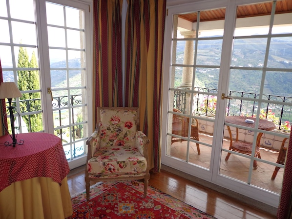Una habitación con un ventanal grande, un sillón floral, una mesita y un tapete estampado.