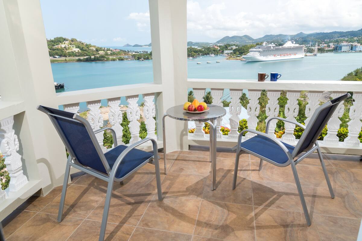 A balcony with two chairs and a small table, overlooking a body of water with a cruise ship and distant hills.