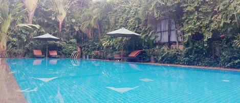 A swimming pool with tiled flooring, surrounded by lush greenery and two umbrellas providing shade.
