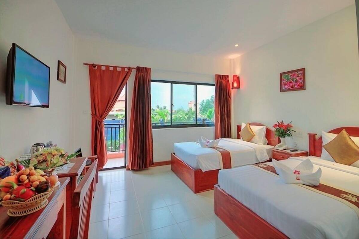 A hotel room with two beds, a TV, a window with curtains, a desk with a basket of fruit, and a painting on the wall.