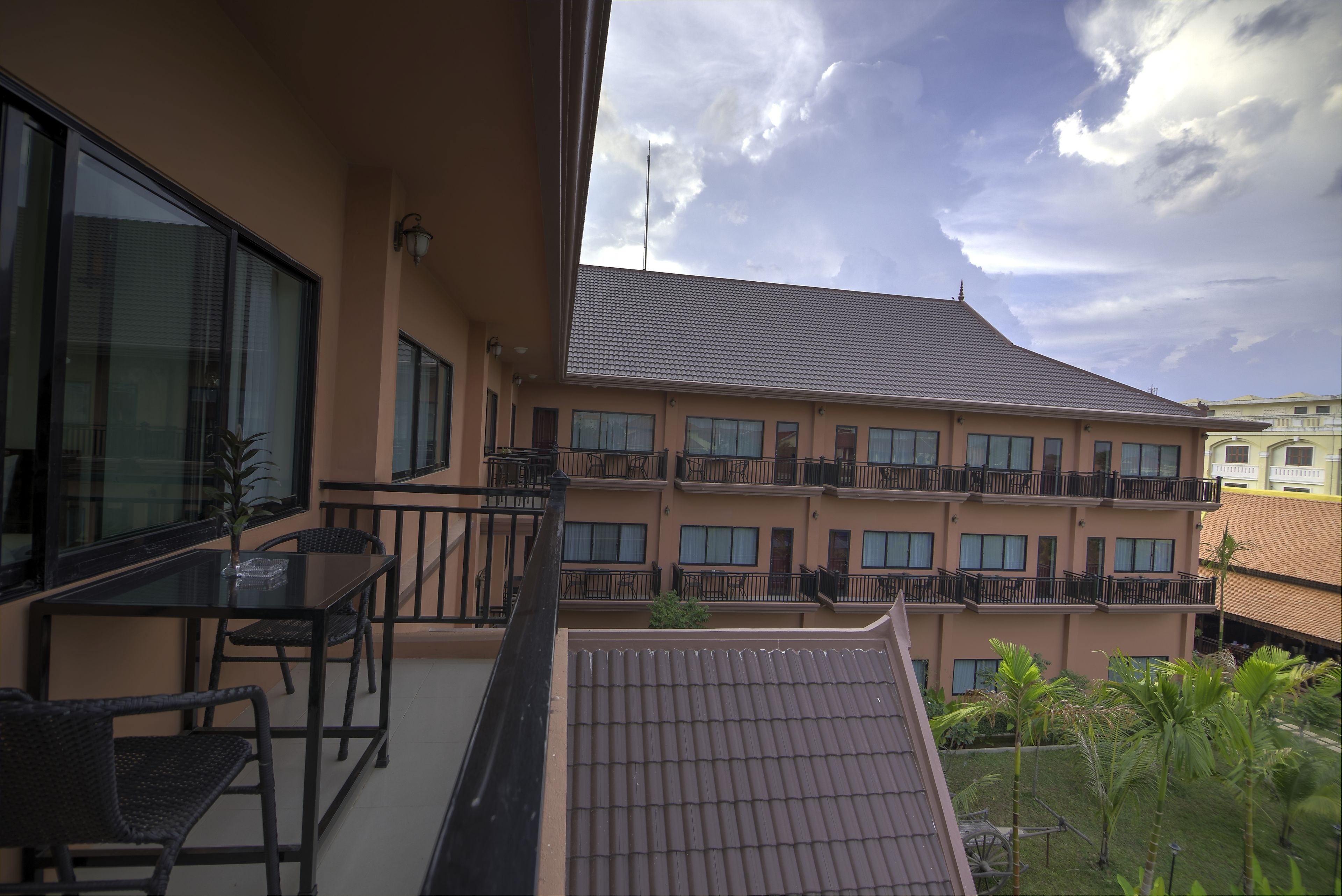A balcony with a glass table and chairs, overlooking a building with multiple balconies and a tiled roof.
