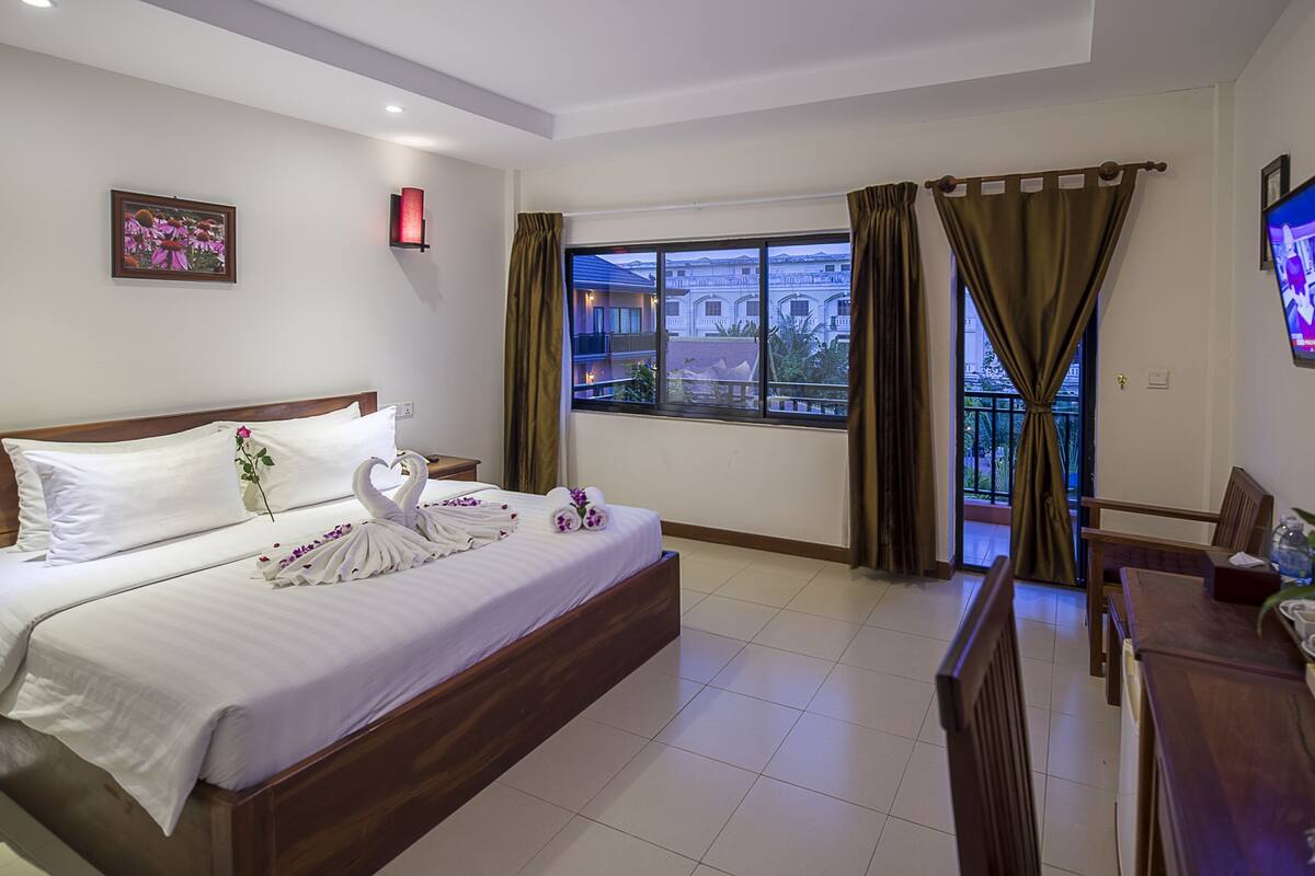 A hotel room with a bed, a TV, a balcony with curtains, and a view of buildings.