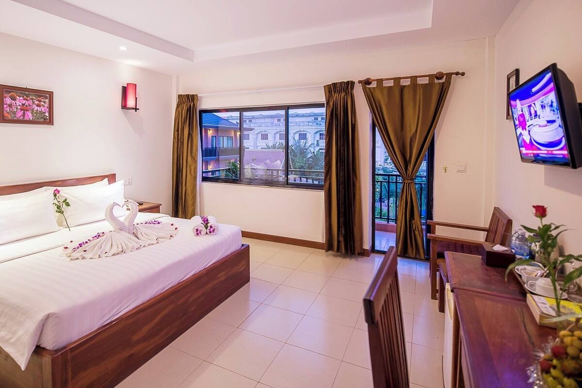 A hotel room with a bed, a TV, a balcony with curtains, and a view of buildings.