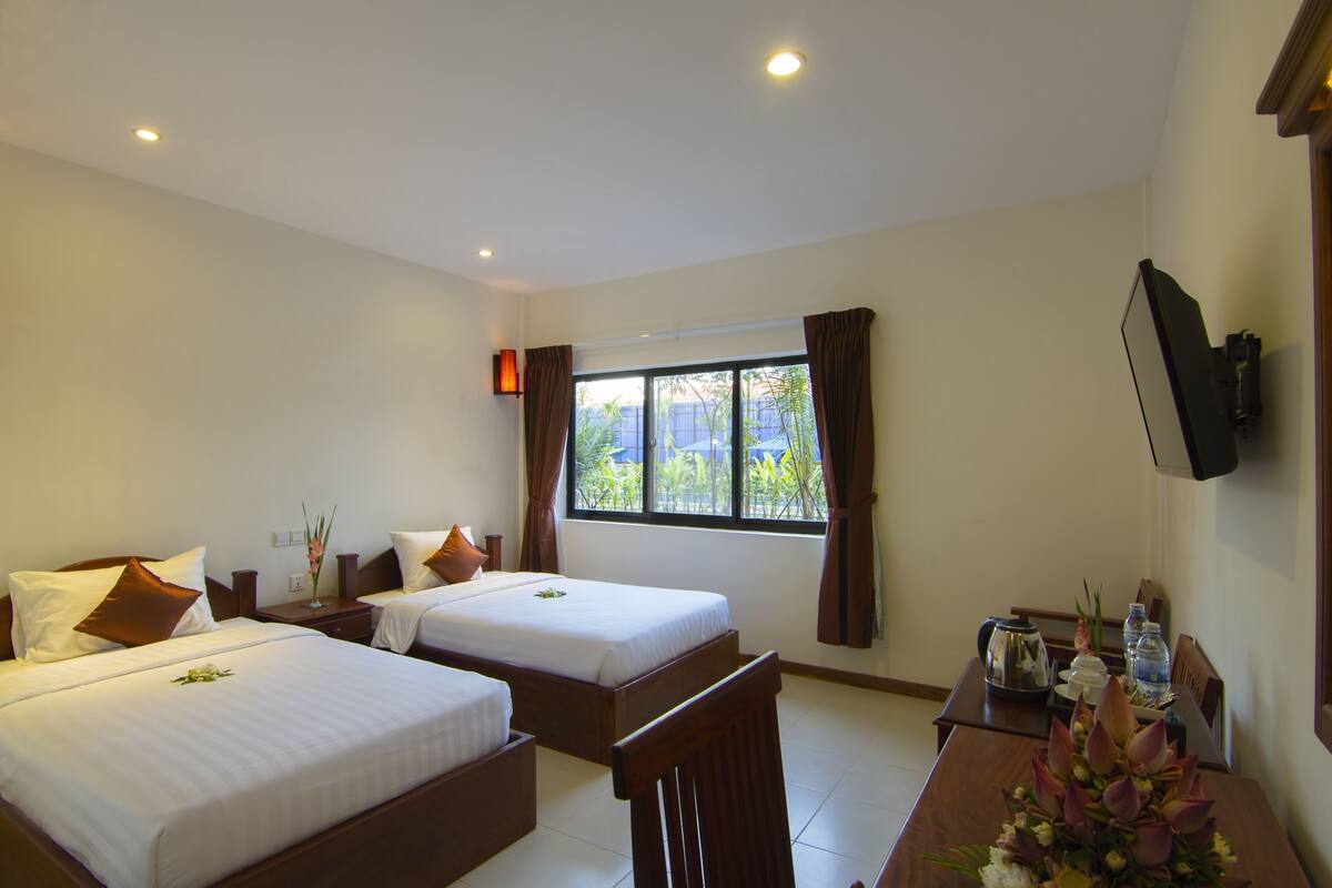 A hotel room with two beds, a desk with a chair, a TV mounted on the wall, and a window with curtains.