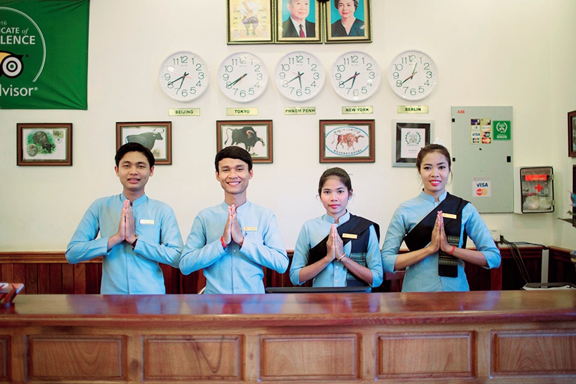 A reception desk with four individuals standing behind it, a wall clock displaying multiple time zones, and framed pictures above the clock.