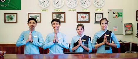 A reception desk with four individuals standing behind it, a wall clock displaying multiple time zones, and framed pictures above the clock.