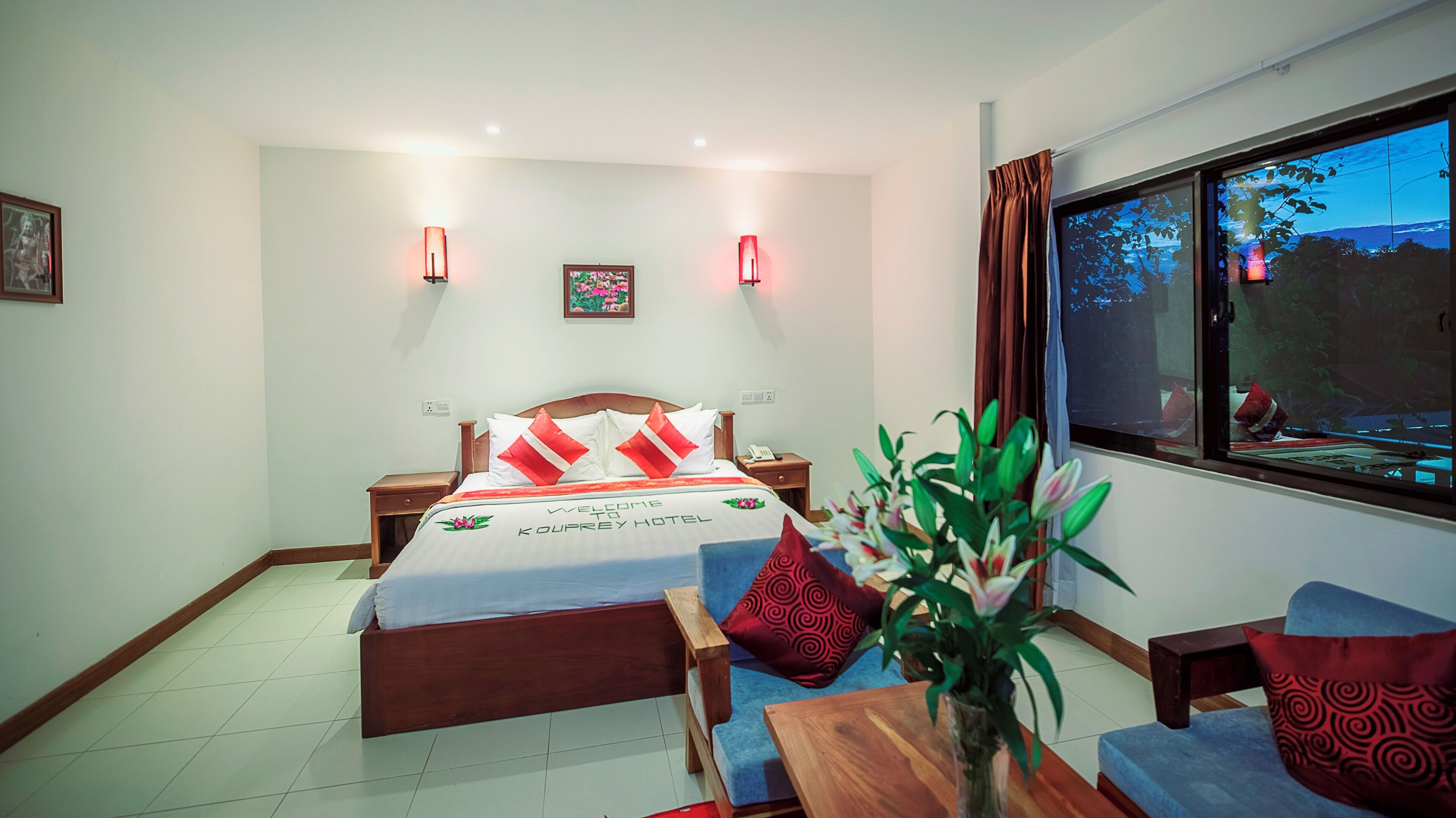 Una habitación de hotel con cama, mesitas de noche, escritorio, silla, una planta en maceta y ventana con vistas.