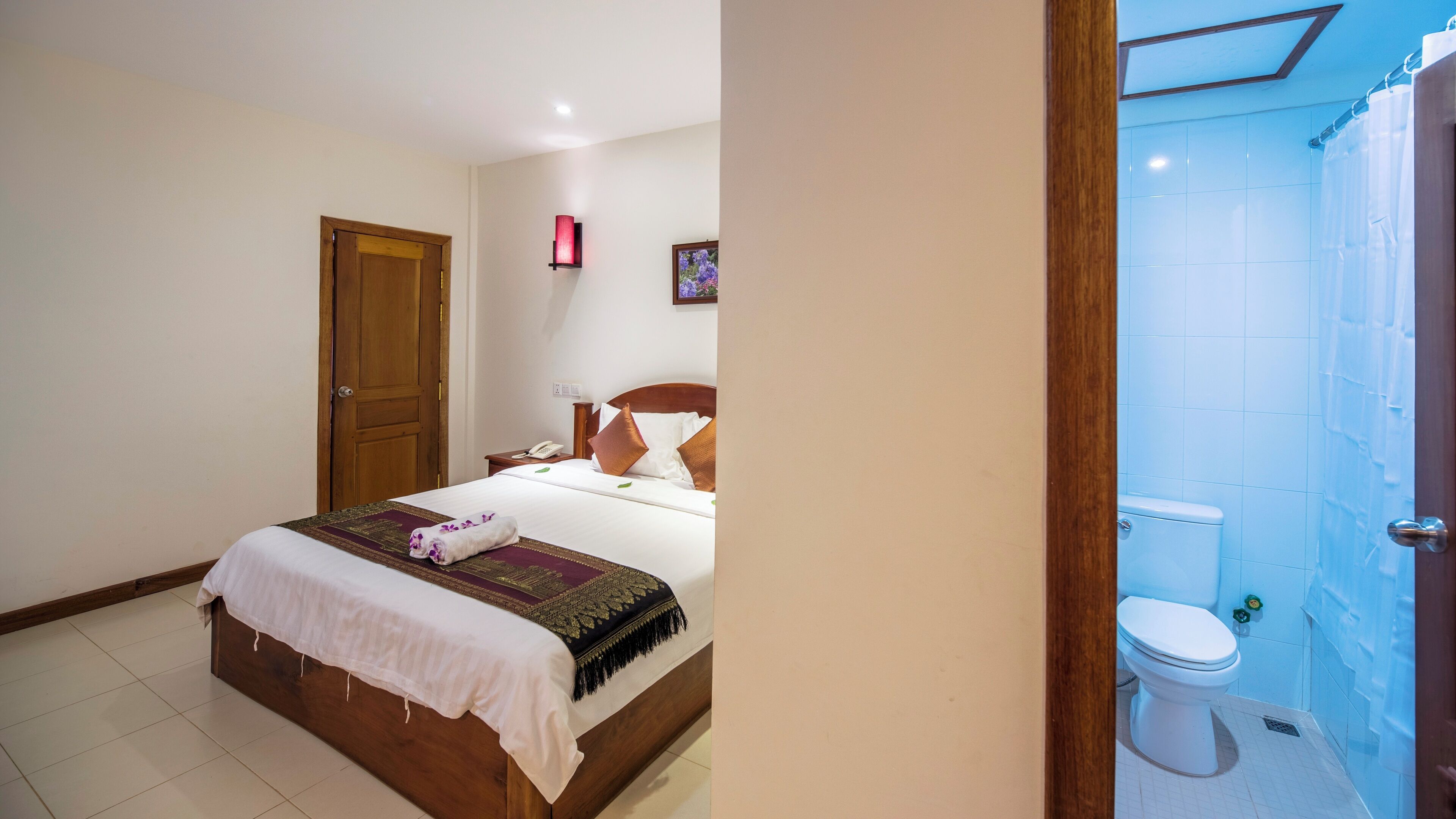 A hotel room with a wooden bed, a bedside table with a lamp, a white toilet, and a shower area.
