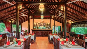 A restaurant with wooden tables and chairs, a bar area, and a view of greenery outside. - Kouprey Hotel (Siem Reap)