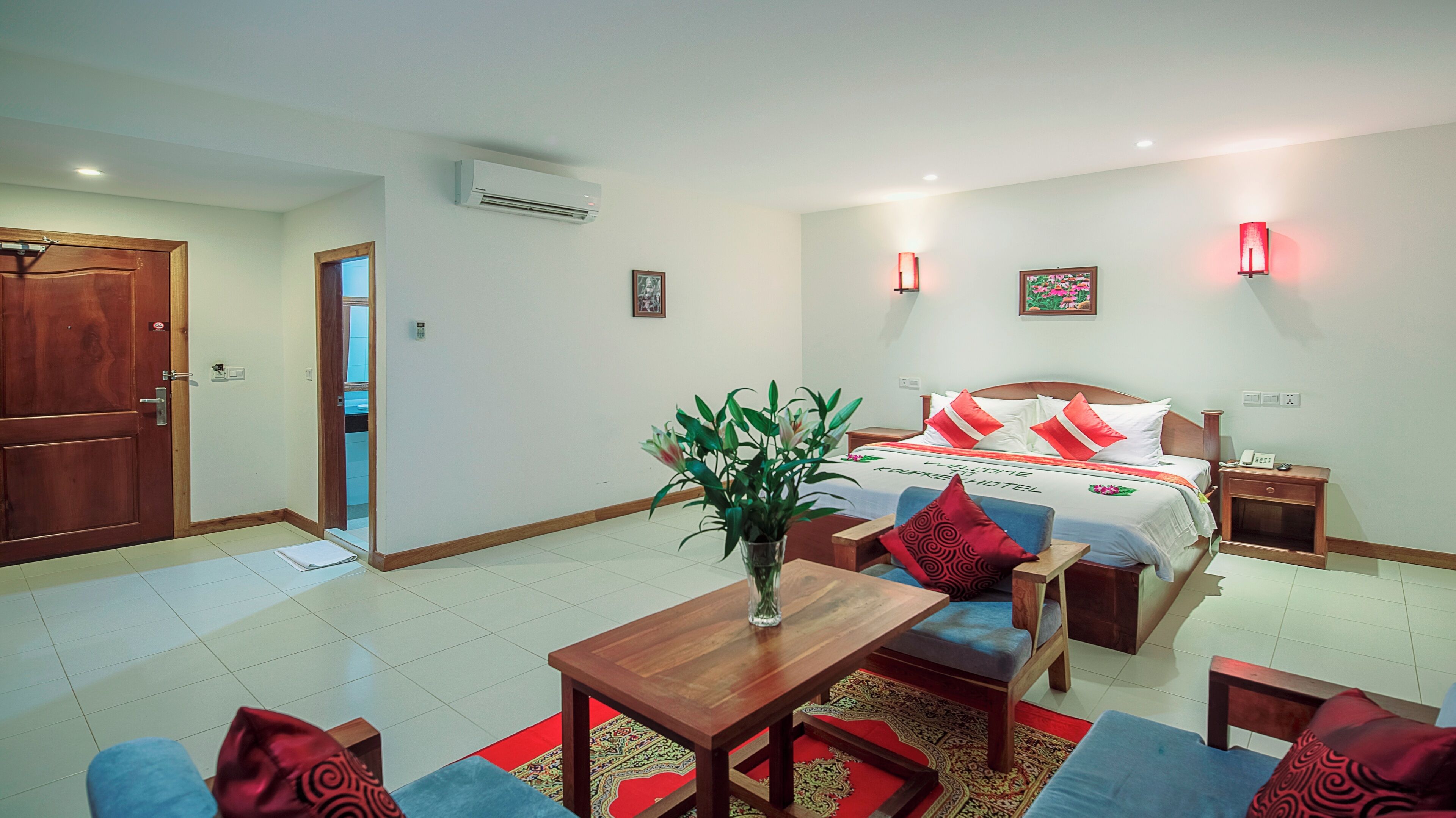 A hotel room with a bed, bedside tables, a desk, a chair, a sofa, a red rug, and a potted plant.