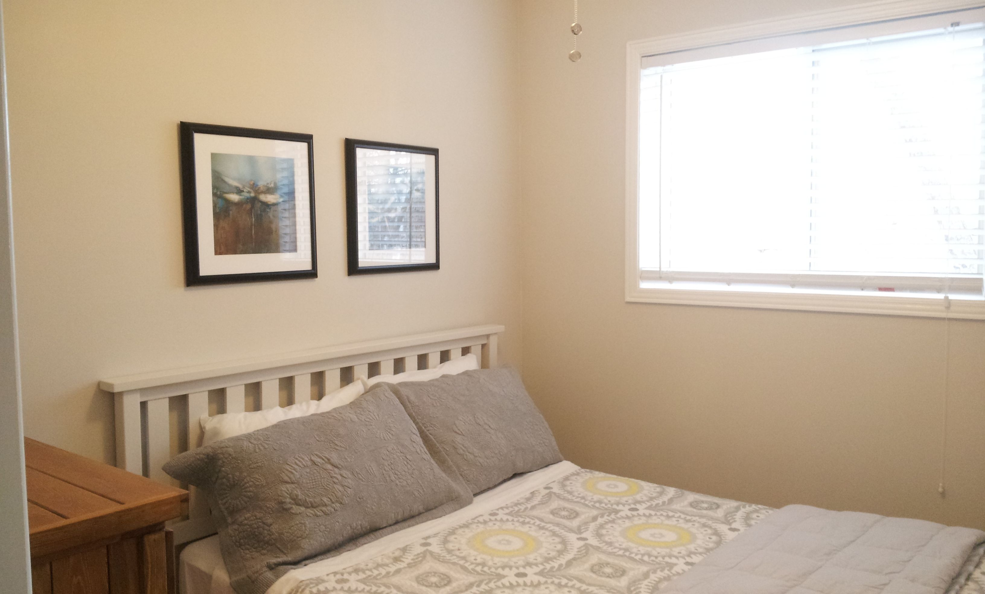 Un dormitorio con cama, mesita de noche, ventana y dos cuadros en la pared.