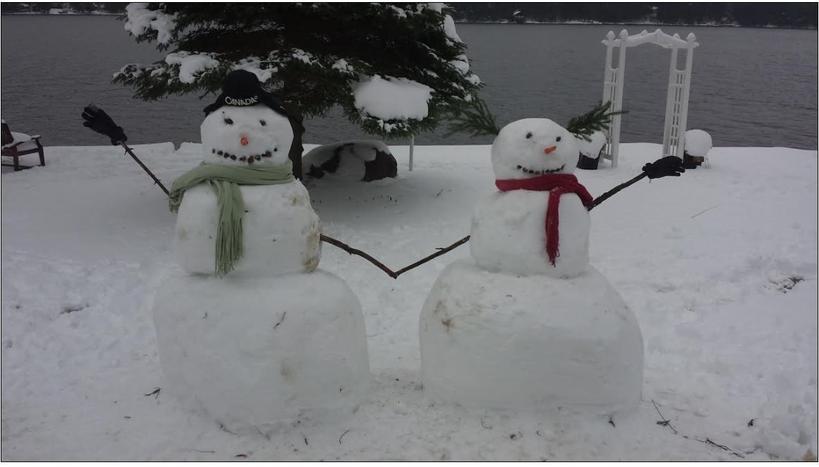 Two snowmen in a snowy outdoor setting with a body of water and a white gazebo in the background.