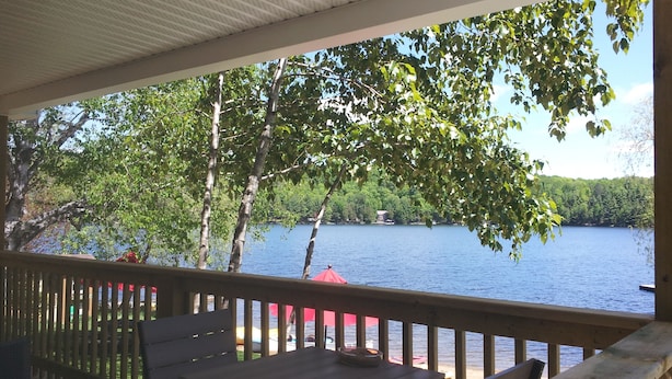 A covered outdoor area with a view of a lake, wooden deck, and a table with a bowl. - Ogopogo Resort (Minden Hills)