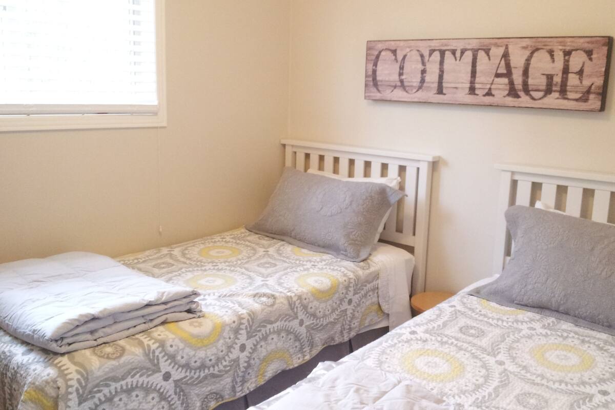 A bedroom with two single beds, each with a patterned bedspread and pillows. A wooden sign with the word "COTTAGE" is mounted on the wall above the beds.