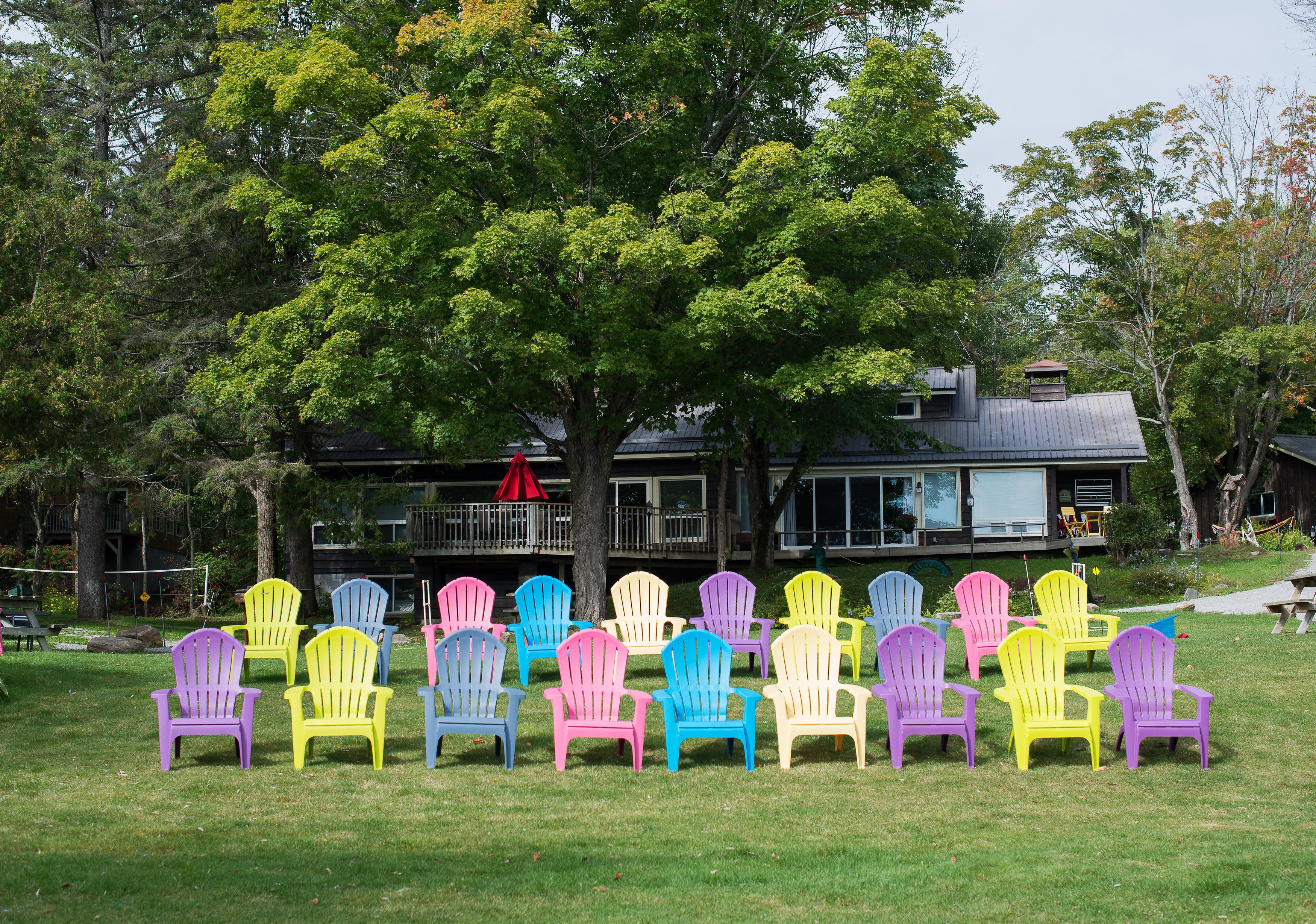 Een rij kleurrijke Adirondack-stoelen, geplaatst in een grasveld, met een grote boom en een gebouw op de achtergrond.