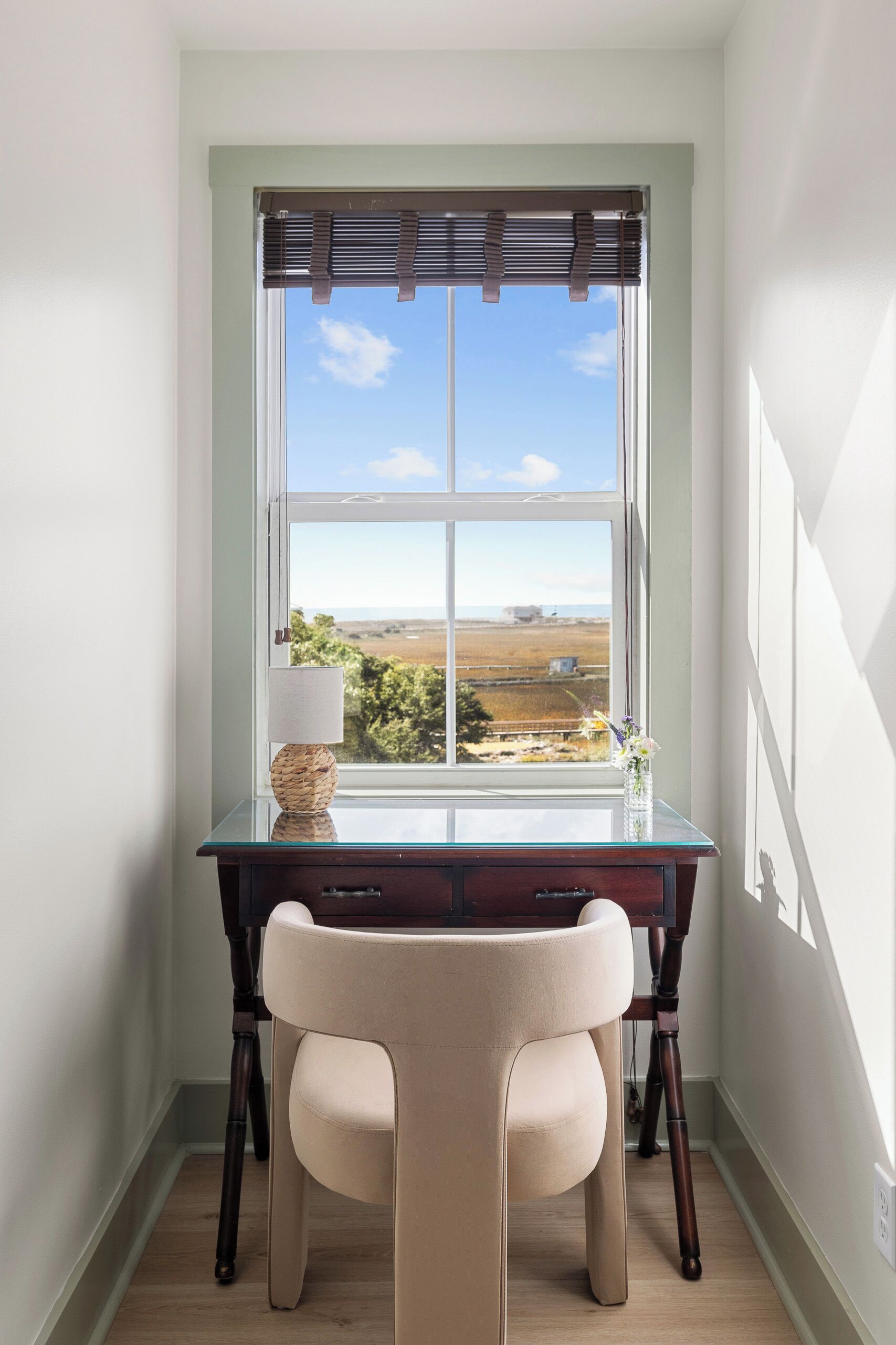 A corner desk with a chair, a lamp, and a view of a landscape through a window with blinds.