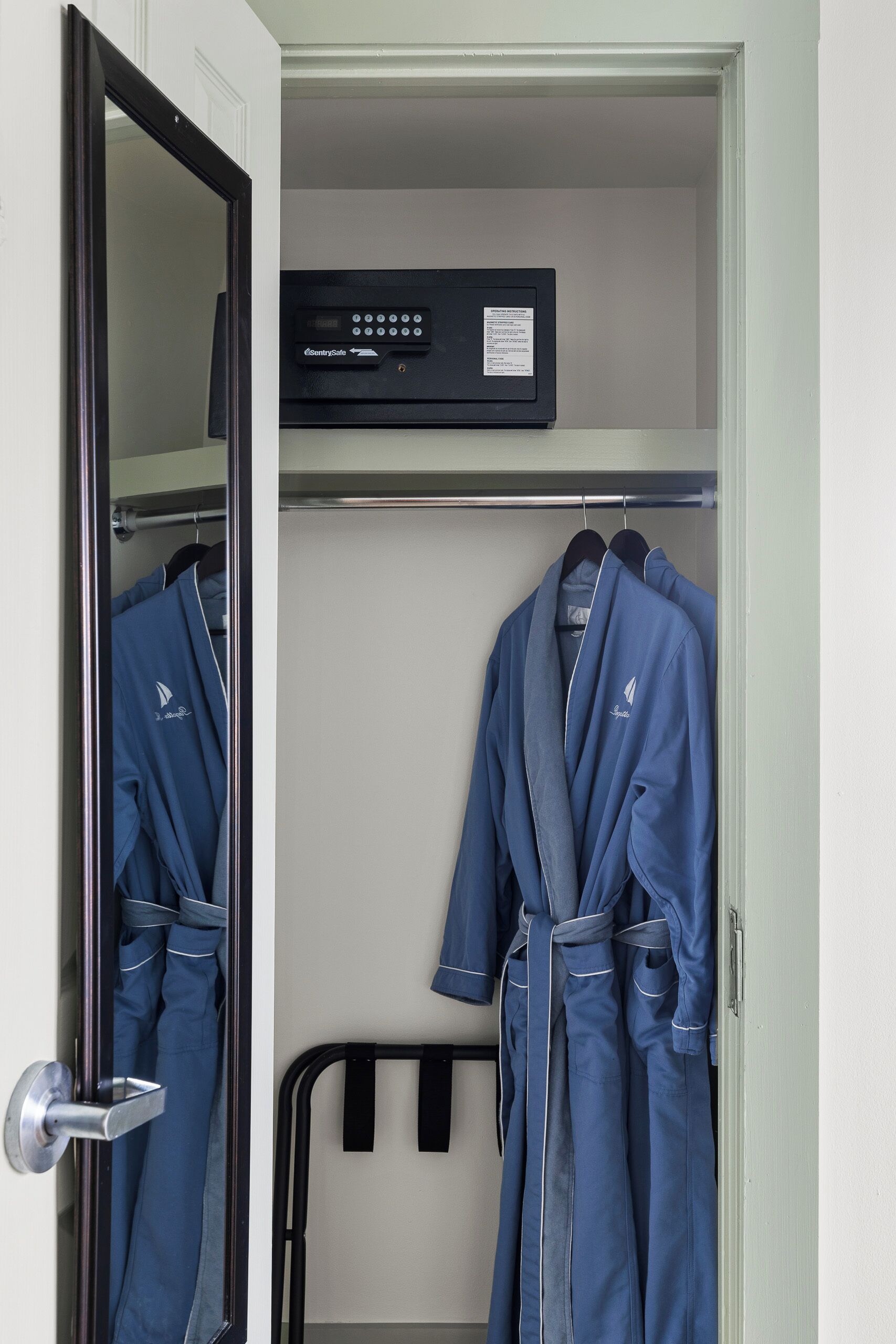 A hotel room with a wardrobe containing blue robes, a black electronic device on the shelf, and a metal rack.