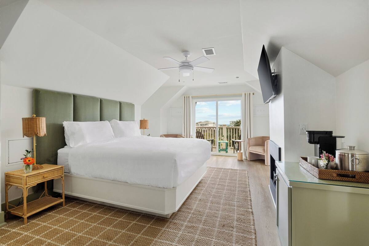 A modern hotel room with a large bed, a flat-screen TV mounted on the wall, a balcony with a view, and a kitchenette.