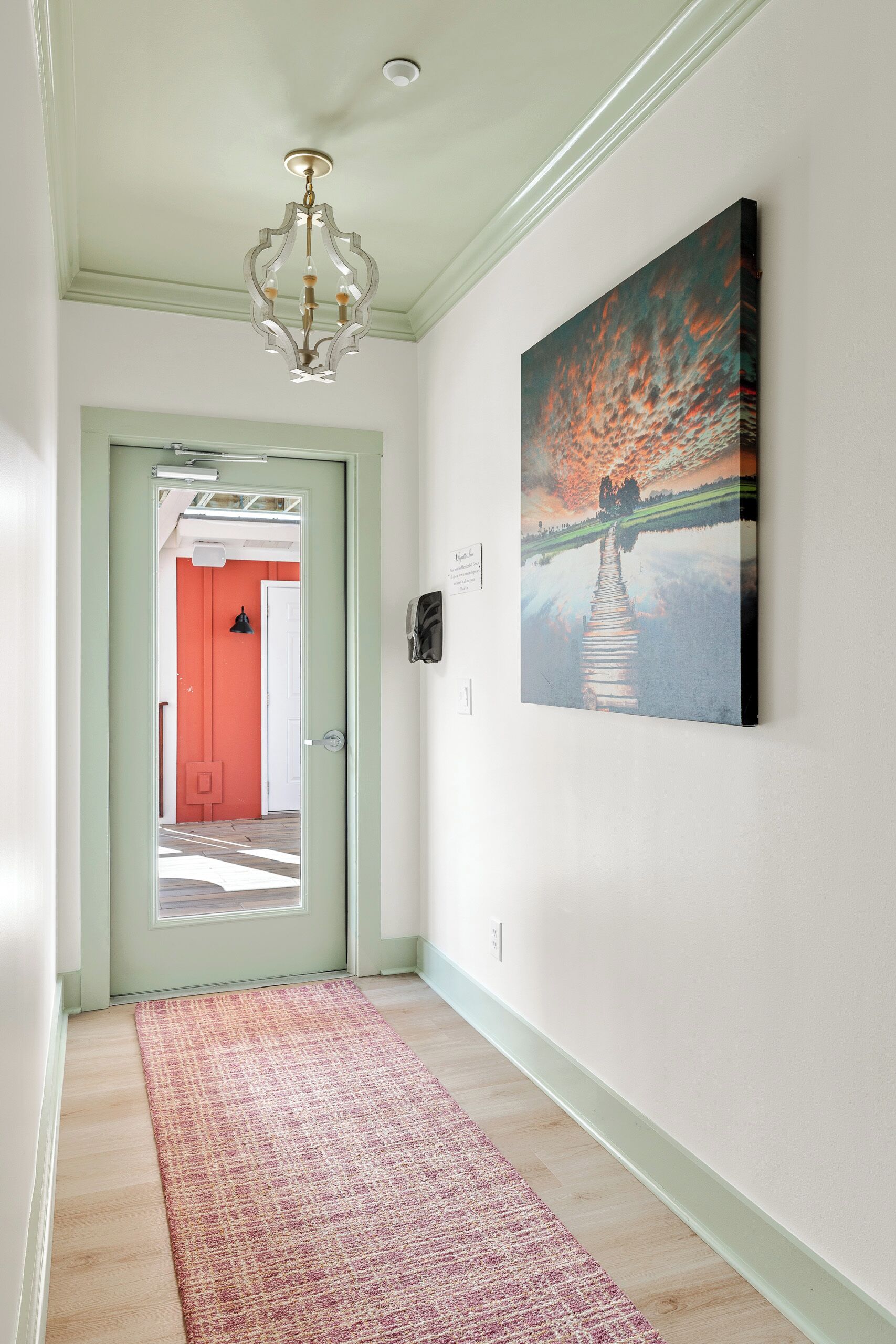 A hallway with a wooden floor, a red door, a chandelier, and a painting of a sunset over a body of water.