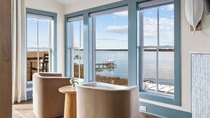 A room with large windows, a wooden table, two chairs, and a view of a dock and water.