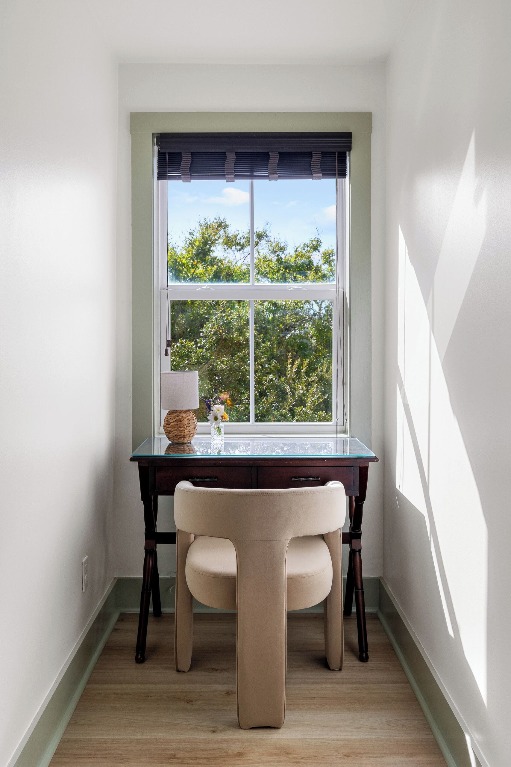 A corner room with a desk and chair, a window with blinds, and a view of trees outside.