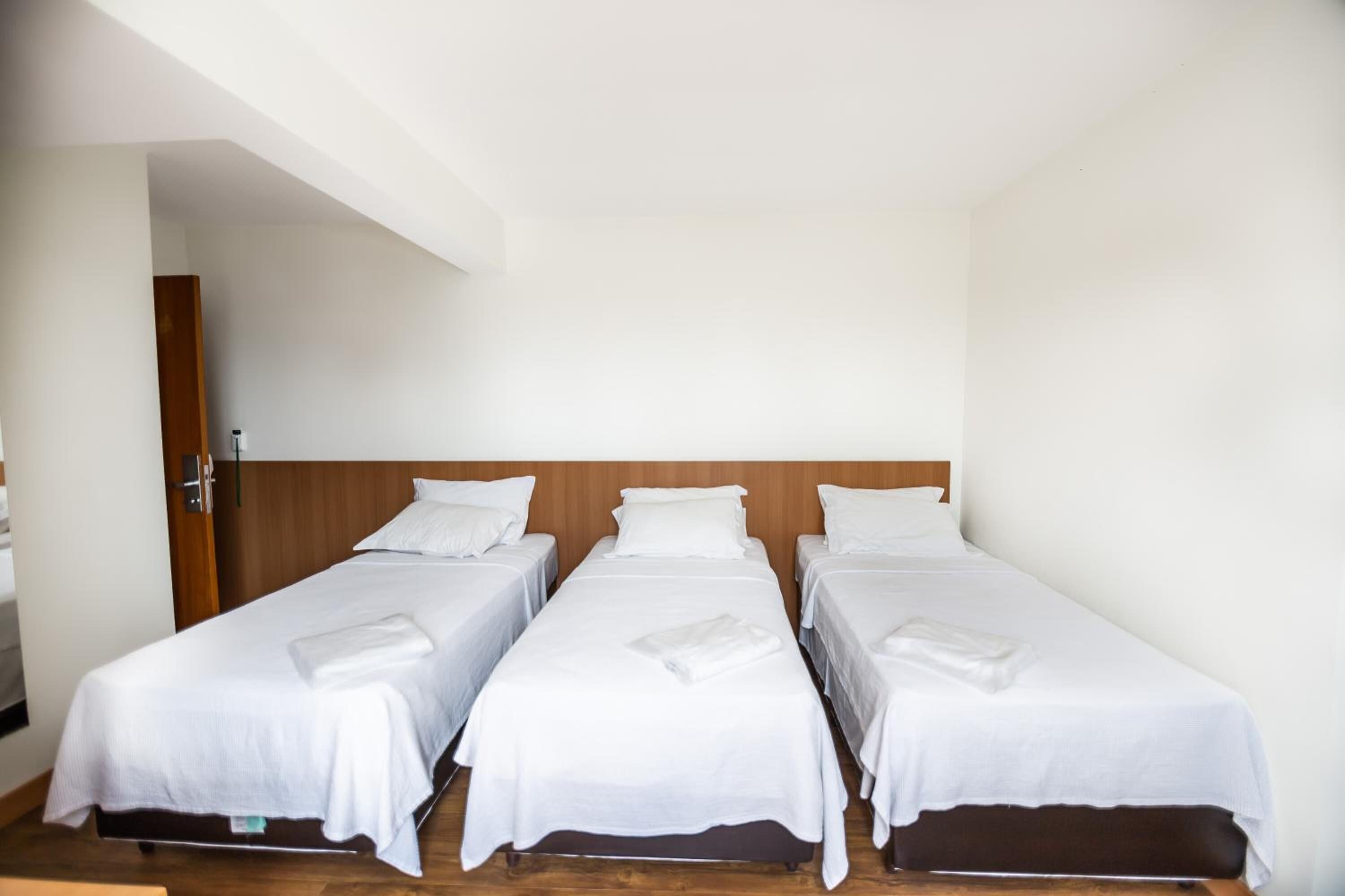 A hotel room with three single beds aligned against a wooden headboard, each with white bedding and a folded towel.