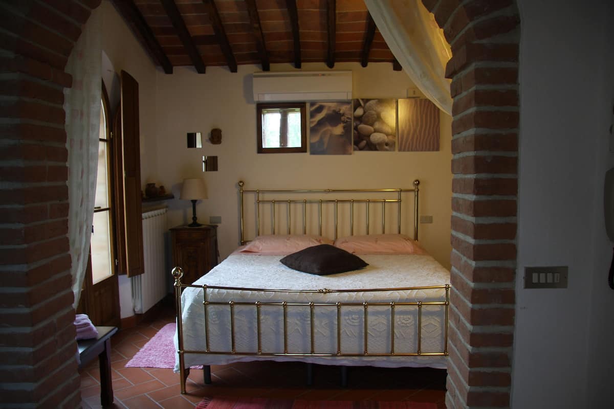 A bedroom with a metal-framed bed, a brick wall, a window, and a wall-mounted air conditioner.