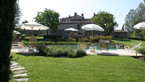 Zona de piscina con sillones de descanso y sombrillas, rodeada de un césped cuidado y edificios de piedra al fondo.