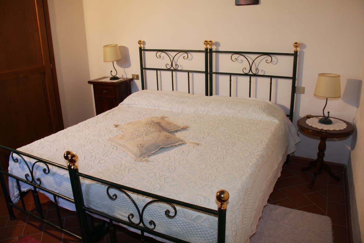 A neatly made bed with a white bedspread and a single pillow, flanked by two bedside tables with lamps, in a room with a wooden door and tiled floor.