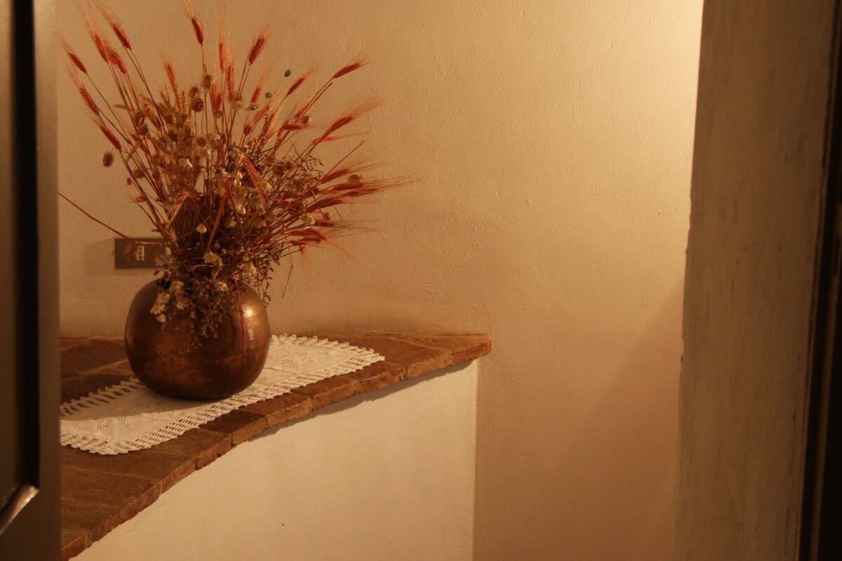 A corner of a room with a vase of dried flowers on a wooden ledge, a wall-mounted light fixture, and a tiled floor.