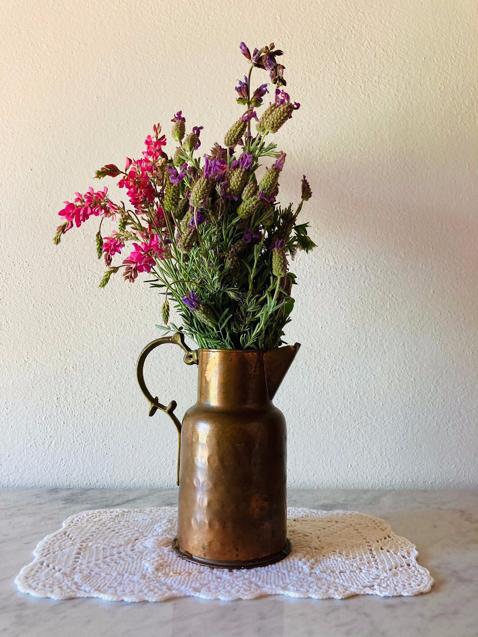 A copper vase with a handle containing pink and purple flowers on a white doily.