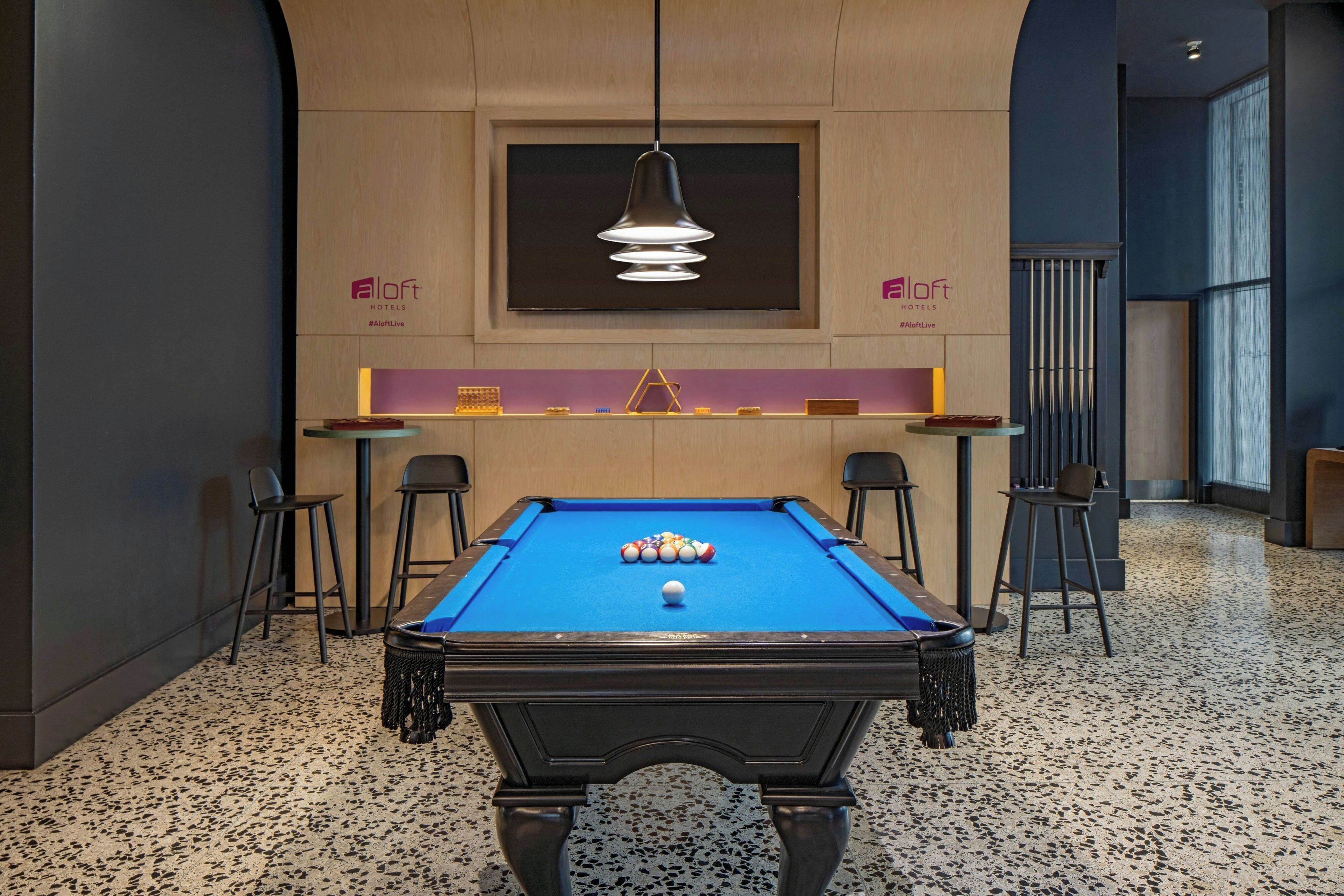 Ein Billardtisch in einem modern gestalteten Raum mit einer Pendelleuchte, einer Bar und einer Sitzecke.