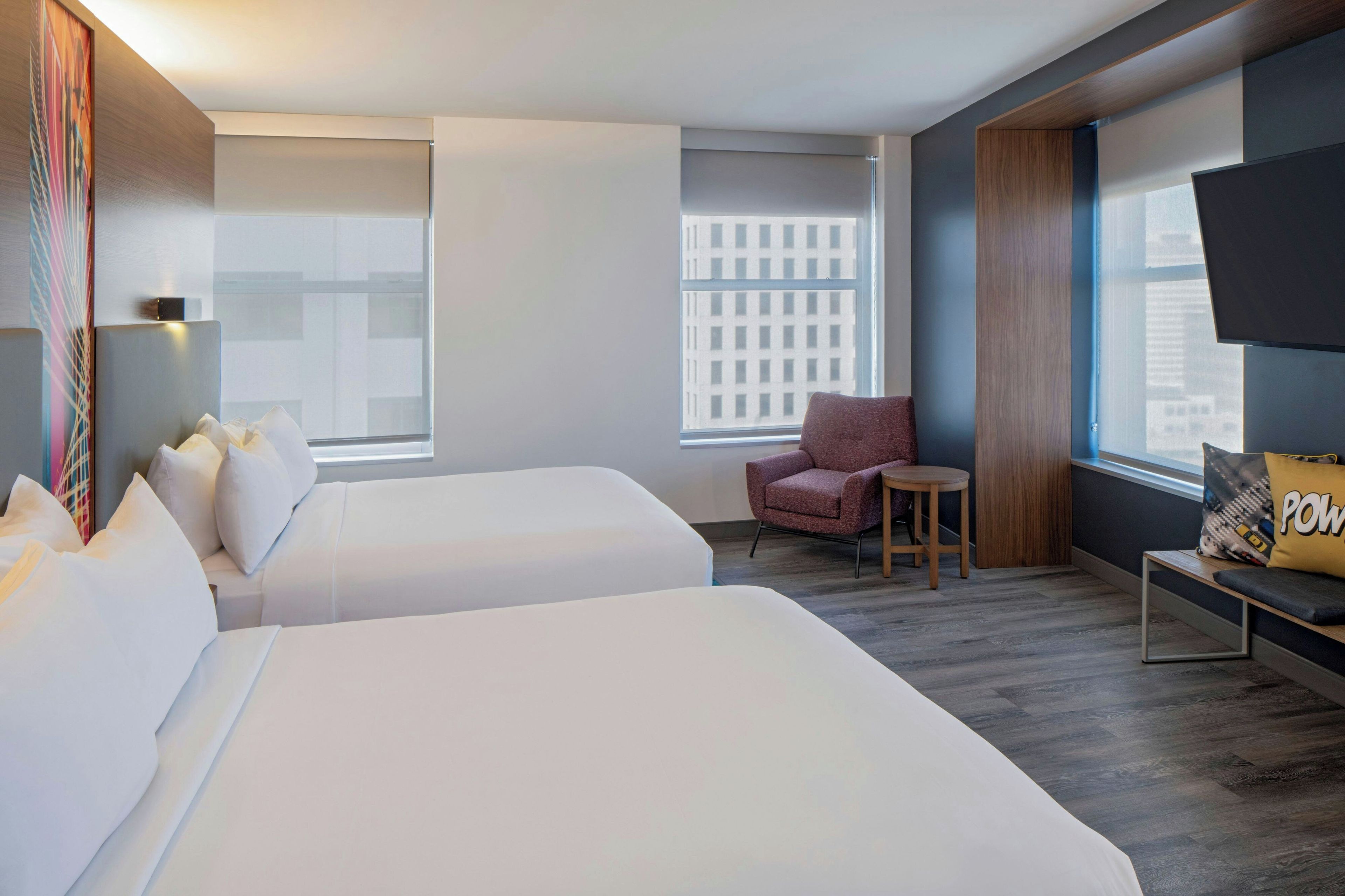 Ein Hotelzimmer mit zwei Betten, einem Sessel, einem kleinen Tisch, einem Fernseher und einem Fenster mit Blick auf die Stadt.