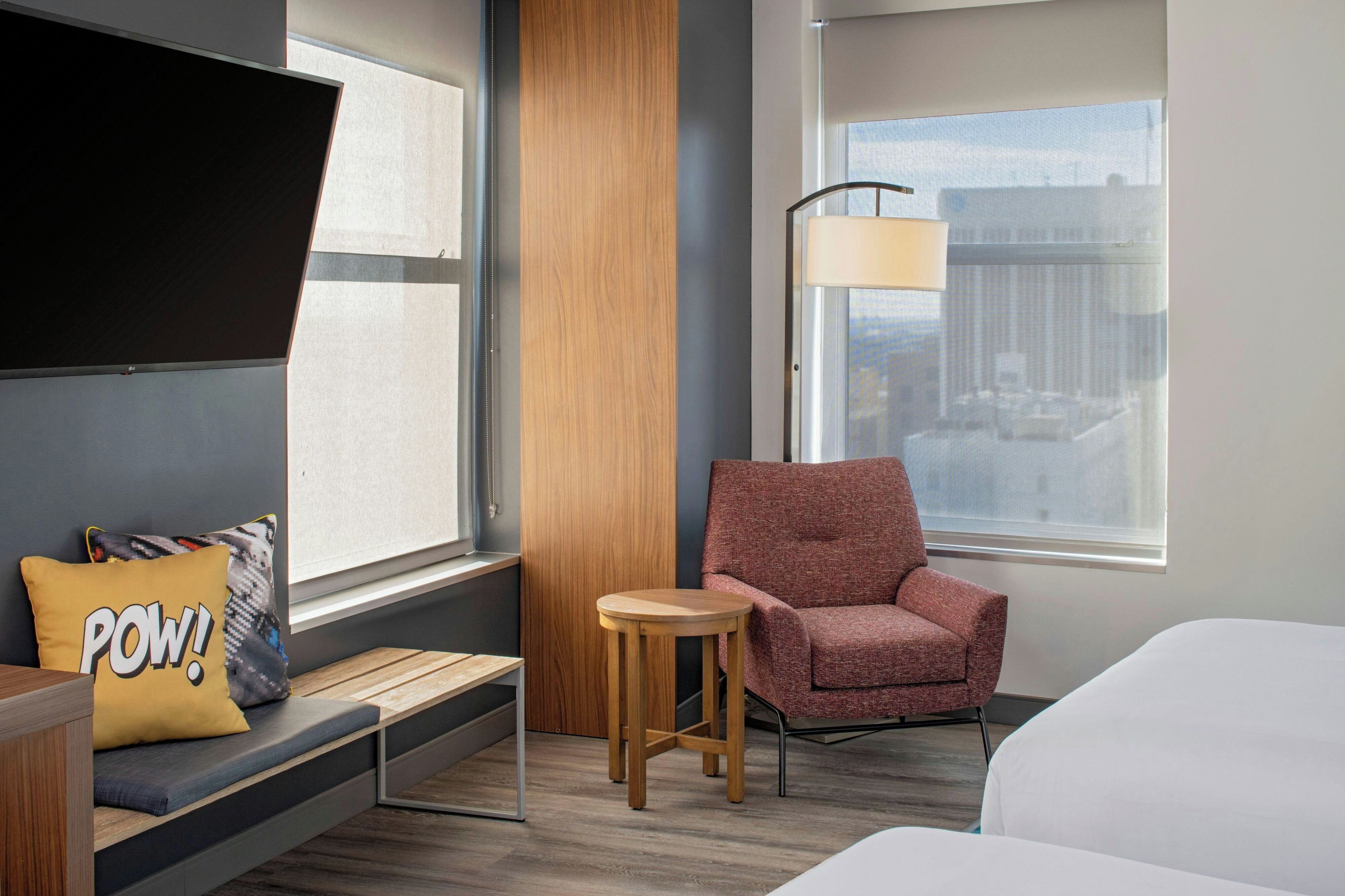 Ein Hotelzimmer mit einem Bett, einer Sitzecke mit einer Bank und einem Sessel, einem kleinen Tisch und einem Fenster mit Blick auf die Stadt.