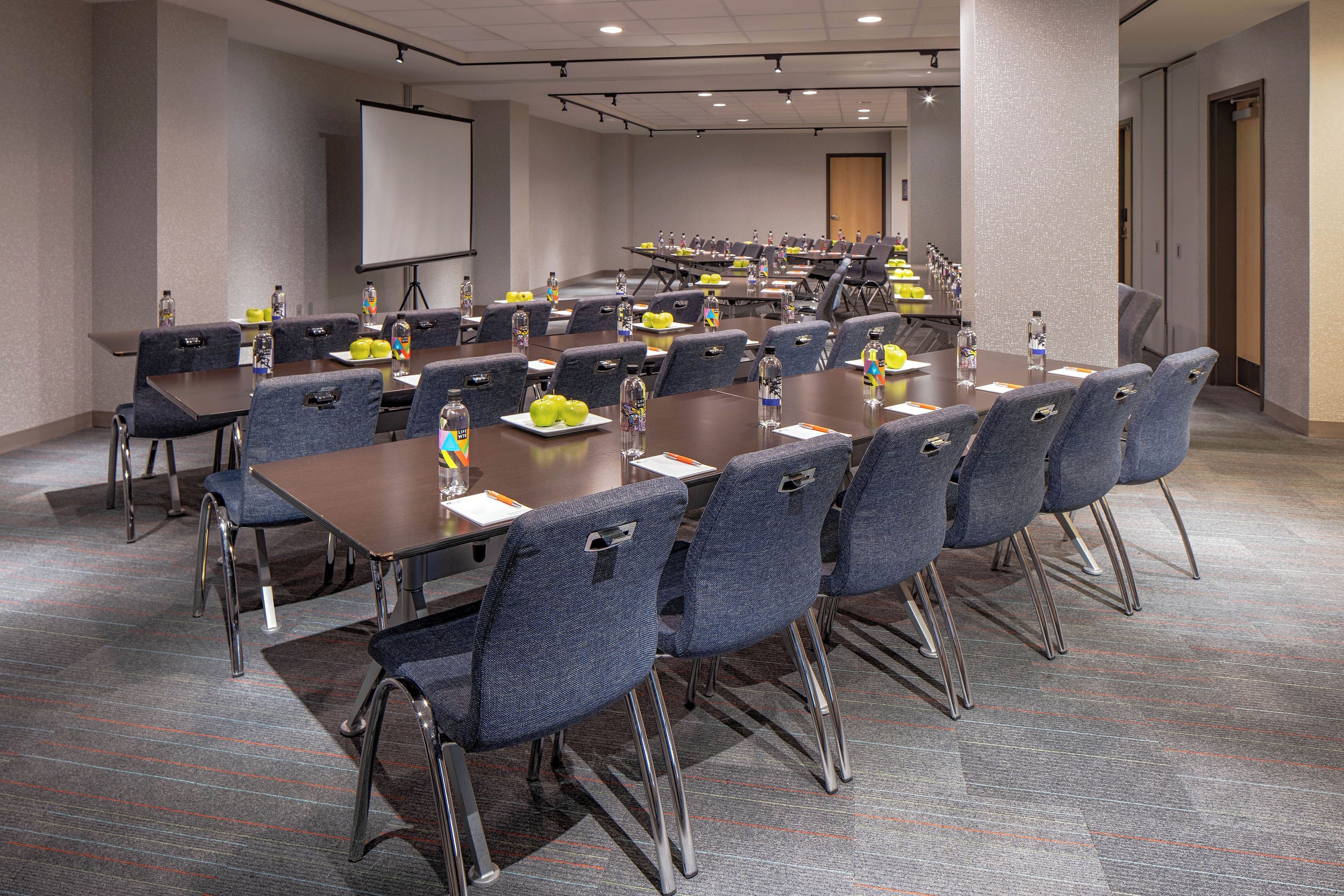 Un salón de conferencias con varias mesas y sillas, equipado con botellas de agua y platos con manzanas.