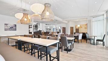 A modern dining area with multiple tables and chairs, pendant lighting, and a view of the kitchen in the background.