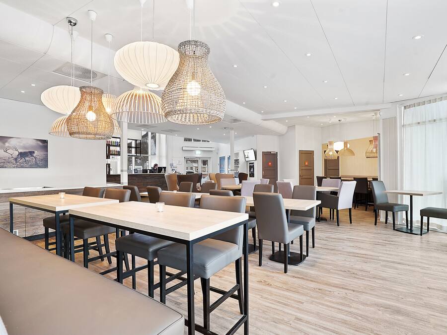 A modern dining area with multiple tables and chairs, pendant lighting, and a view of the kitchen in the background.