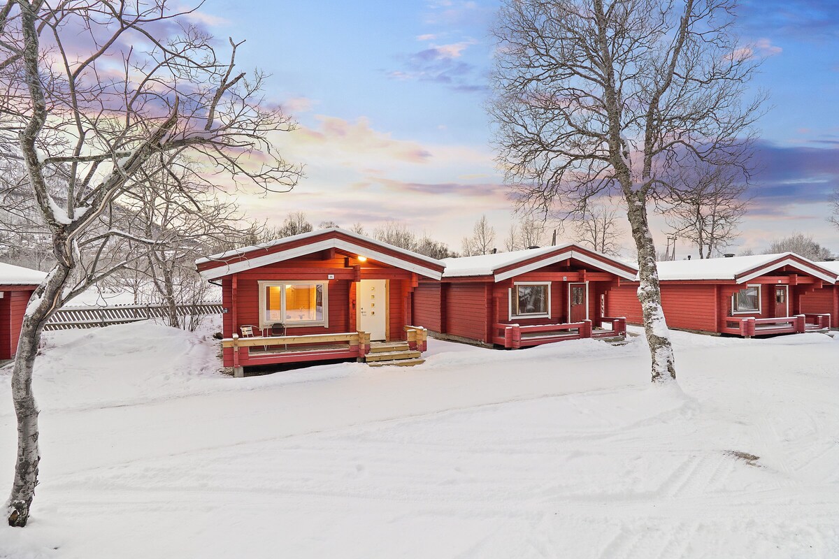 Una hilera de cabañas rojas sobre un terreno cubierto de nieve y árboles sin hojas, indicando un paisaje invernal.