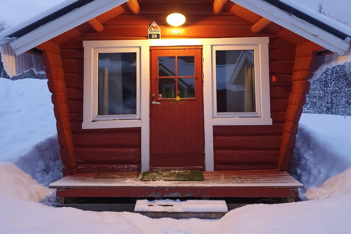 Eine kleine, hölzerne Hütte mit roter Tür und zwei Fenstern, umgeben von Schnee.
