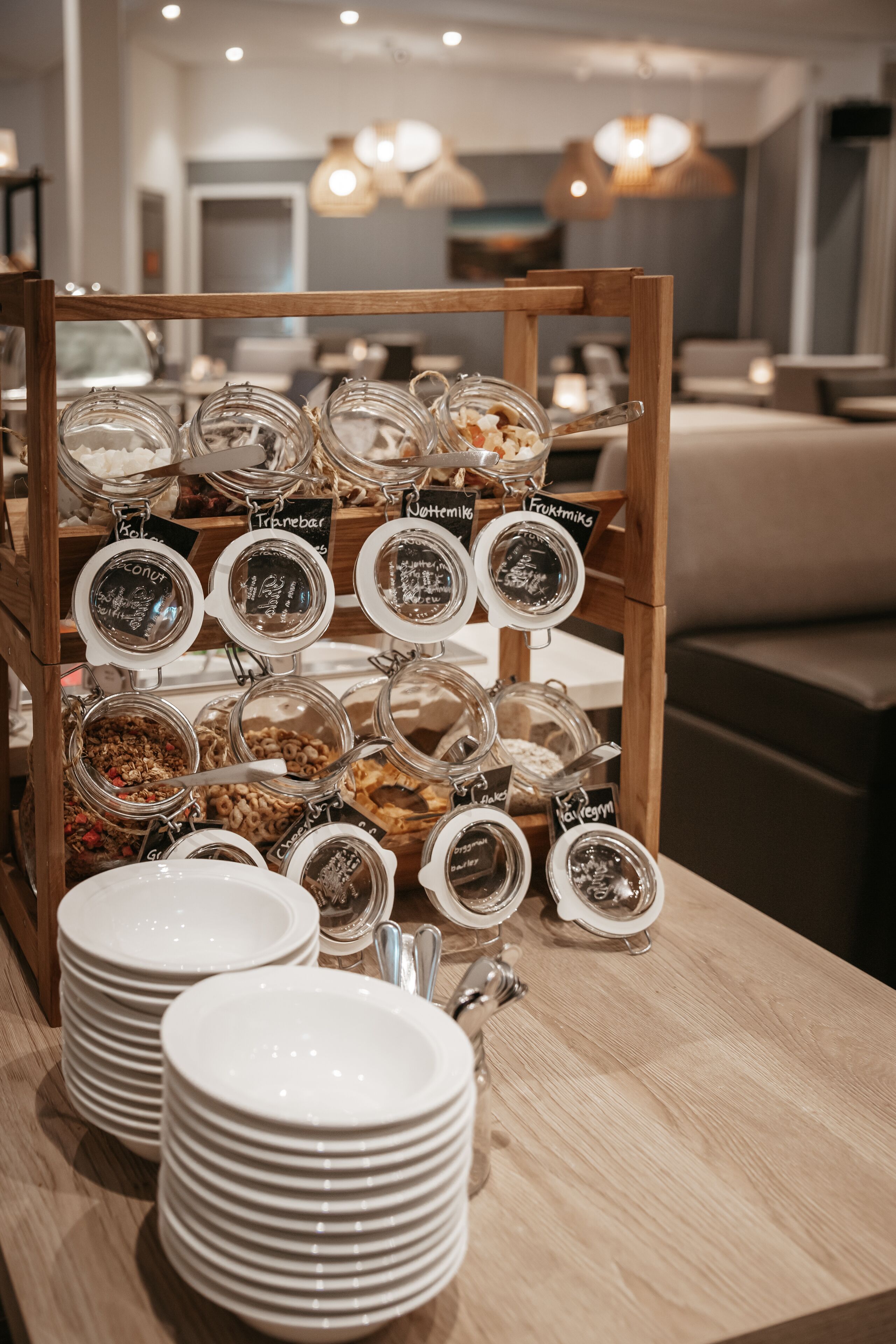 Una presentación de alimentos en frascos con etiquetas, platos apilados y un soporte de madera en un comedor.
