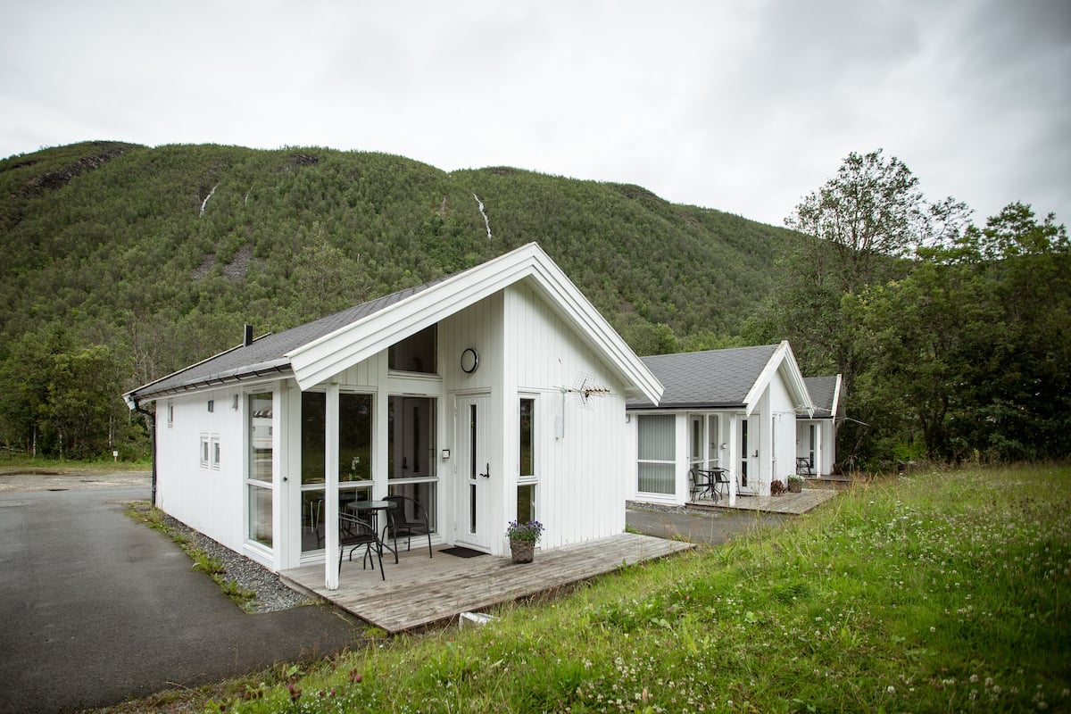 Eine weiße Holzhütte mit Veranda und Essbereich, vor einer bewaldeten Hügelkulisse gelegen.